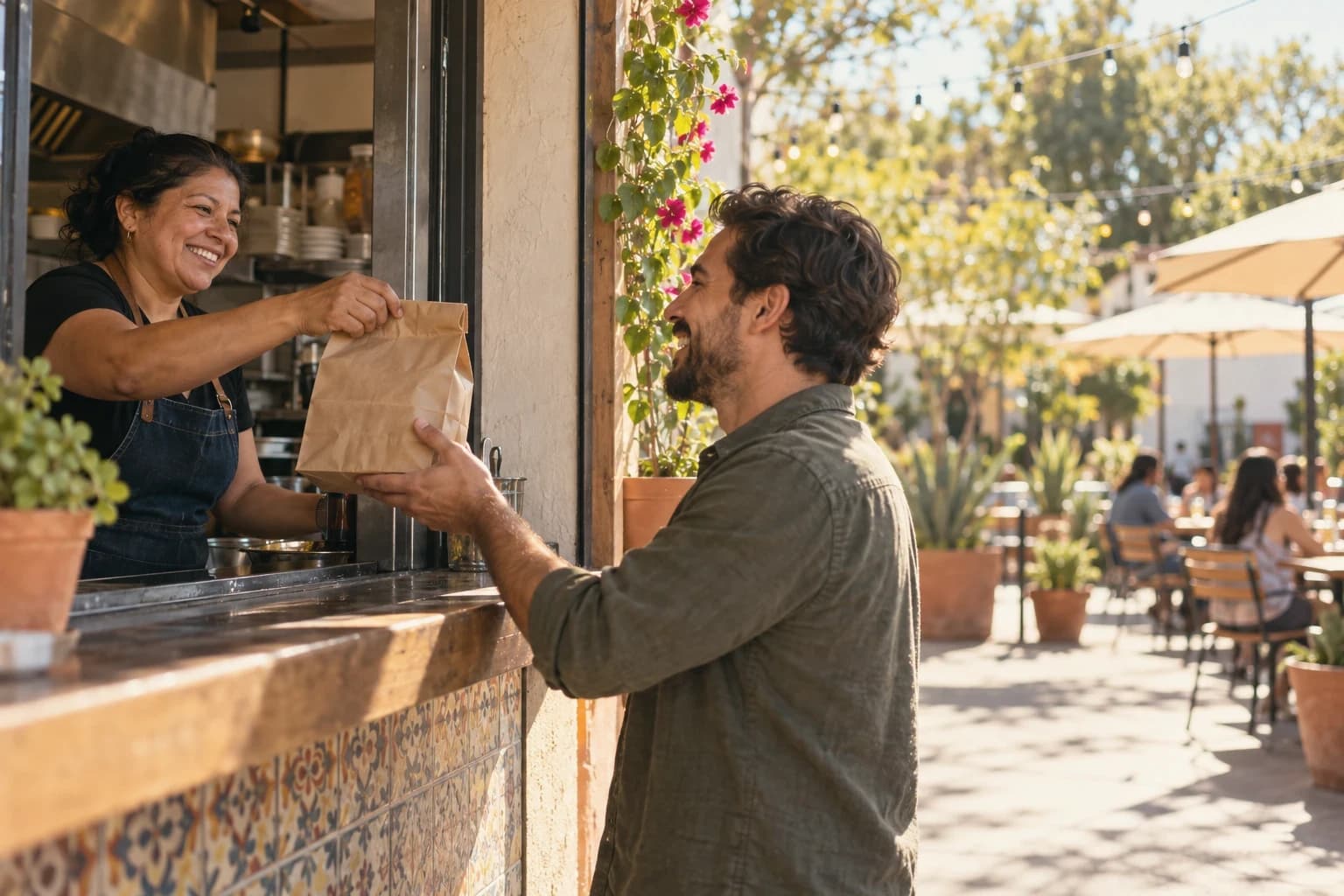 A taqueria owner passes a paper bag through the service window to a smiling customer, a sunny patio with string lights behind them.
